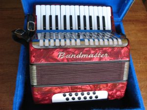 Bandmaster Piano Accordion in marbled red. 26 keys – two octaves, 6 bass and 6 chord buttons. In original case. Leather straps and body in good order. Recently cleaned inside and keys adjusted. Plays well. Made in East Germany